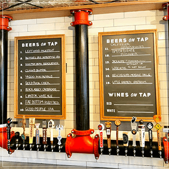Red and black beer towers hanging from the ceiling with multiple taps and 2 wooden menus hanging on a white wall.